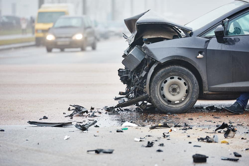 A gray car with severe front-end damage after a collision, debris scattered around.